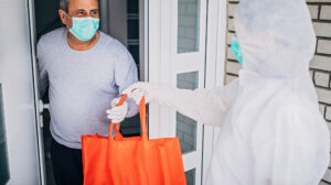 infection control worker dressed in protective clothing hands over a bag of groceries to a man wearing a mask
