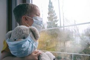Child wearing mask with a teddy staring out a window