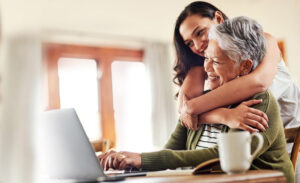 young woman embracing older woman as they sit at a kitchen table using a computer