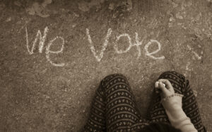 Young person writing "we vote" in chalk